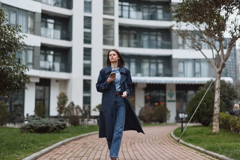 One woman walks along a brick path in an urban courtyard, wearing a dark trench over denim, her gaze ahead, a calm confident stride, soft breeze in her hair.