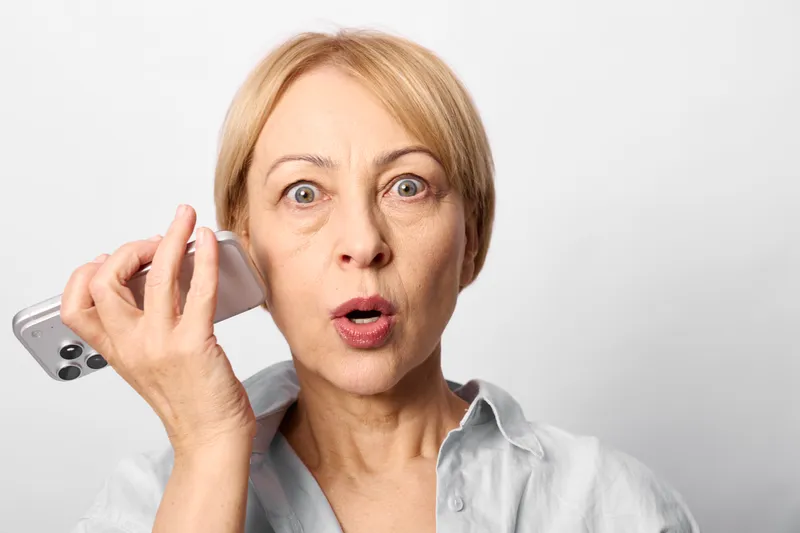 Surprised woman holding a phone to her ear, wide eyes and open mouth, shocked expression. Bright background. Concept of call, communication, reaction, female.