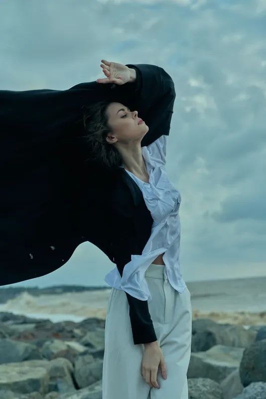 Woman with flowing cape on a windswept beach, dramatic pose under a cloudy sky, fashion forward and free, outdoor scene capturing motion and resilience in coastal landscape