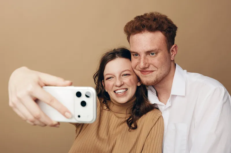 Smiling young couple takes a selfie together on a smartphone. They look happy and close, posing against a plain beige background. Concept of friendship and modern technology.
