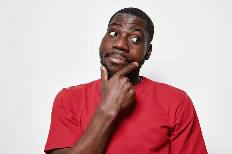 African American man with dark skin wearing red shirt looking thoughtful with hand on chin against white background. Portrait of pensive young black male in casual clothing.