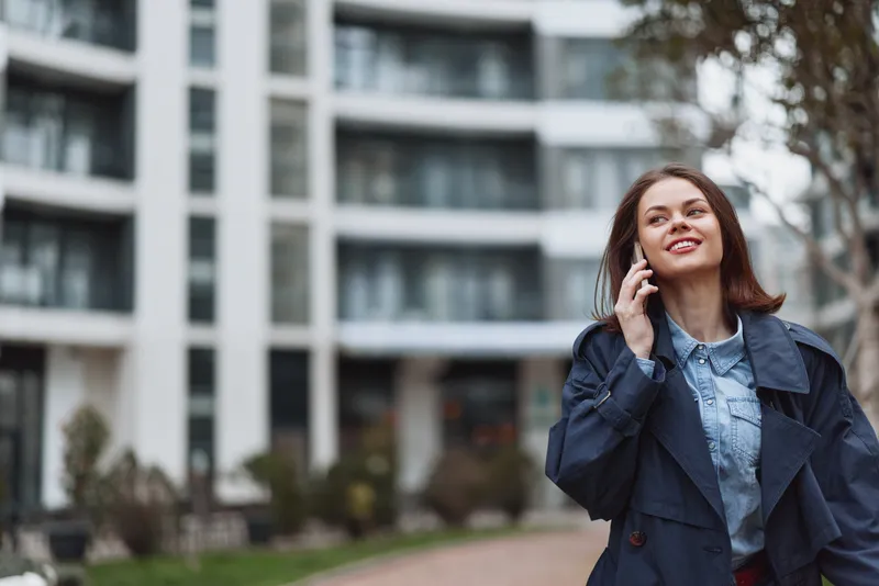 A young woman in a blue coat talks on a smartphone outdoors near modern apartment buildings on a cloudy day. She smiles while walking on a paved path with greenery around.