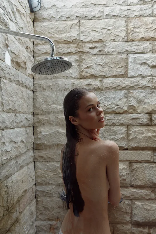 naked woman under rain shower with wet long hair stands in stone shower stall looking back in natural light, skin wet and relaxed atmosphere, wellness concept