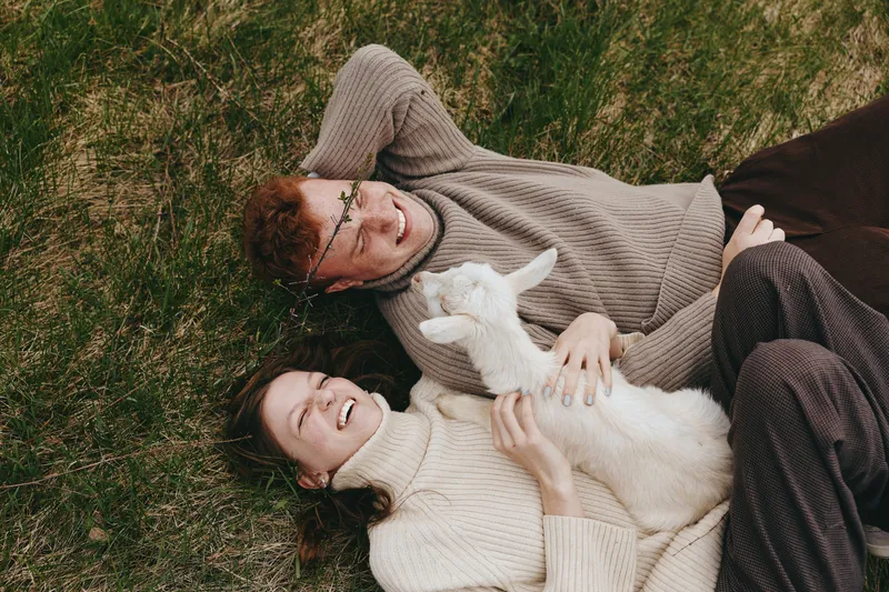 A relaxed couple lies on grass with a playful goat kid between them, capturing a warm countryside moment of lifestyle, affection, and animal companionship under bright daytime light.