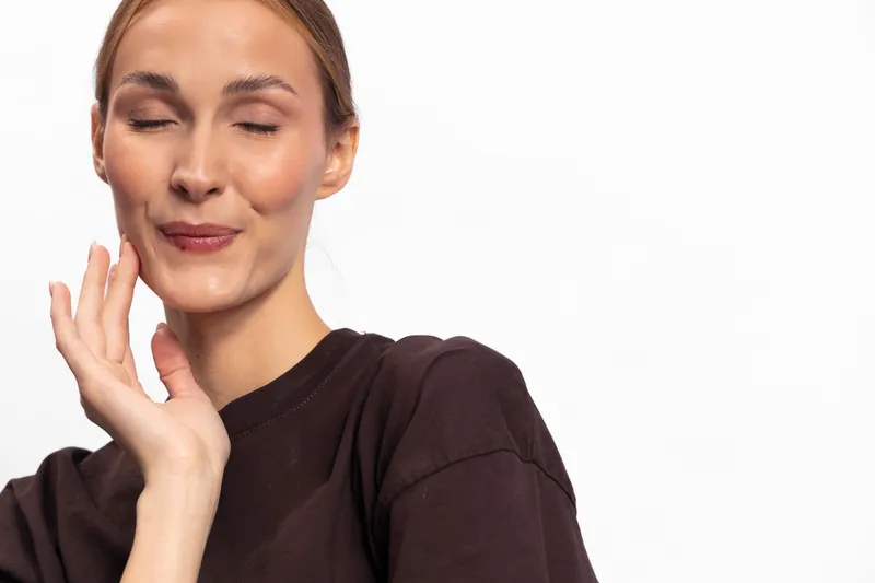 Smiling woman with closed eyes and relaxed face touches cheek gently. Studio portrait on white background. Concept of calm, self-care, beauty, and peaceful moment.