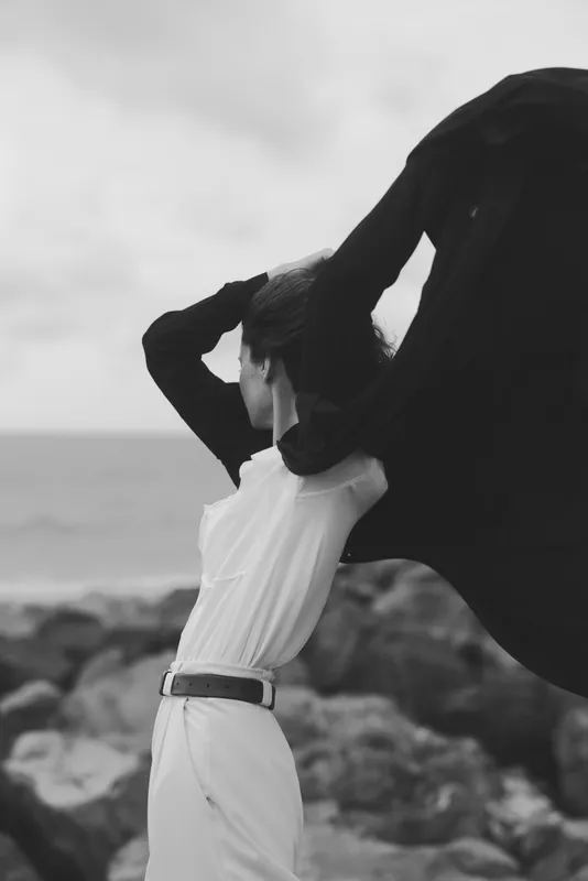 White dress on a windy beach, cape sweeping behind, figure facing the sea, dramatic motion in a monochrome scene, bold fashion photography with natural light