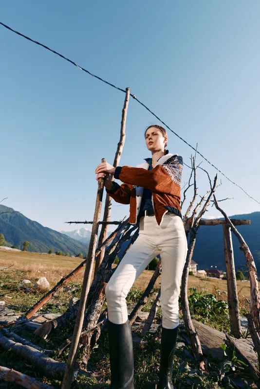 woman with stick in field by mountains wearing boots and patterned sweater, standing confident in rural outdoors scene with clear blue sky, meadow, rustic fence and scenic valley view