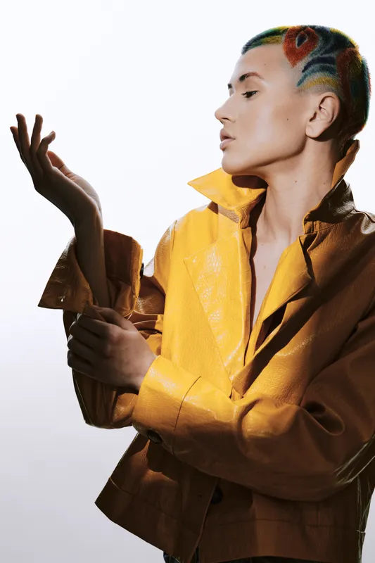 artistic woman with colorful shaved hairstyle wearing a yellow jacket poses thoughtfully against a white background in a studio setting