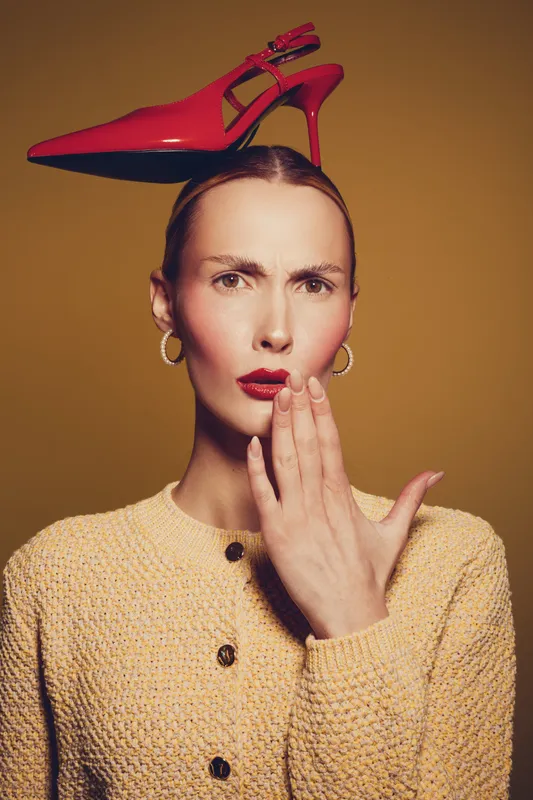 Woman with red high heel shoe balanced on head looks surprised with hand near mouth. Studio portrait with warm lighting and beige background. Concept of fashion and unusual style.