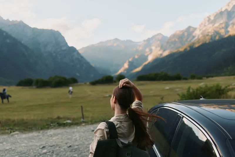 Woman traveler standing beside a car in a mountain valley, backpack over her shoulder, gazing toward distant peaks. Outdoor lifestyle, exploration, and calm nature.