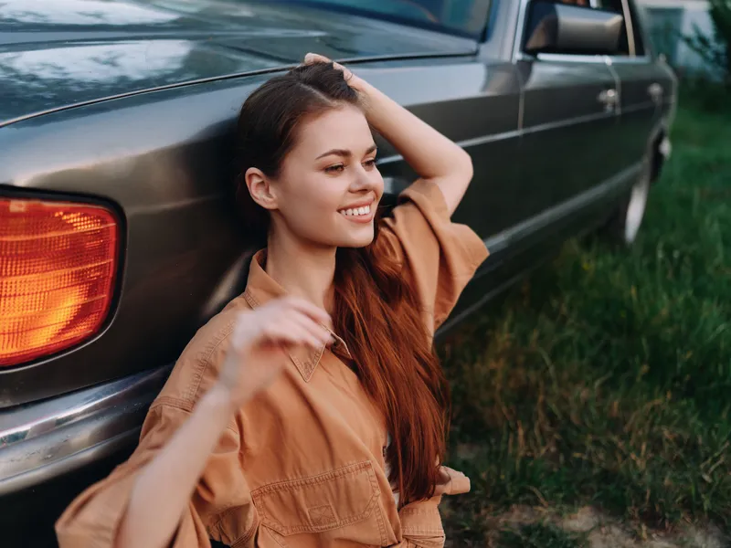 Young woman in casual attire smiling near vintage car, enjoying sunny day in nature, showcasing carefree lifestyle and confidence.