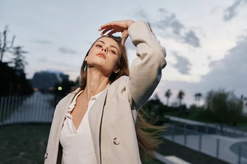 Woman in beige coat touching hair confidently outdoors during cloudy sunset with soft natural light and urban blurred background, casual fashion portrait