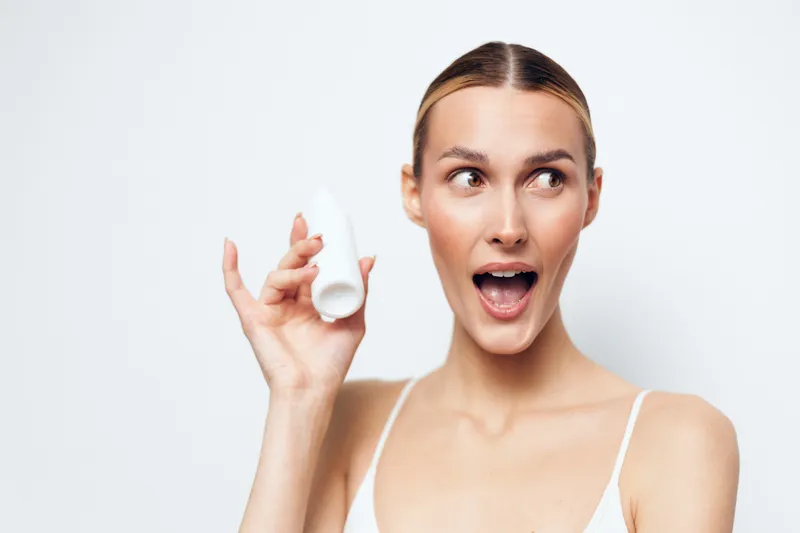 Woman applying skincare beauty cream with cotton pad, surprised open mouth expression, close portrait on white studio background, natural skin, soft light, clean hygiene