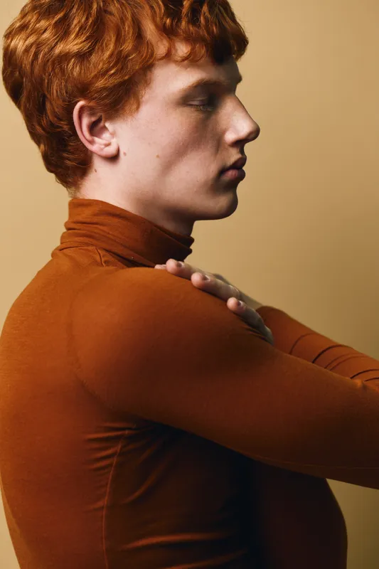 Profile of young man with red curly hair and fair skin wearing brown turtleneck sweater touching his shoulder against neutral background, side view closeup portrait.