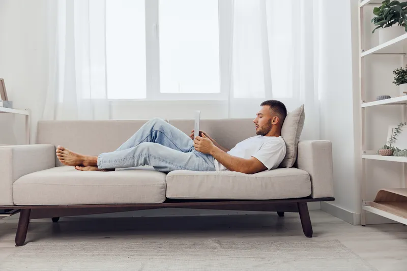 Relaxed man reading a book on a cozy sofa in a bright, modern living room. The natural light enhances the tranquil atmosphere and serene mood.