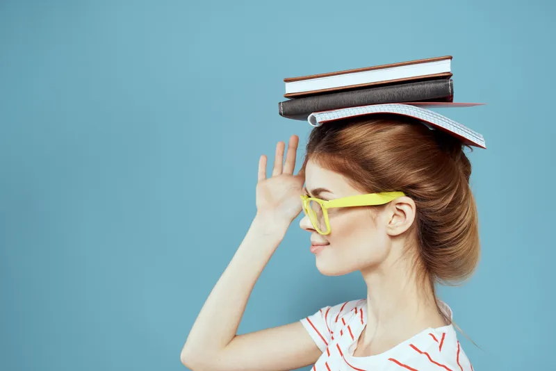Студентка держит книги на голове и в жёлтых очках на синем фоне, обрезанный вид