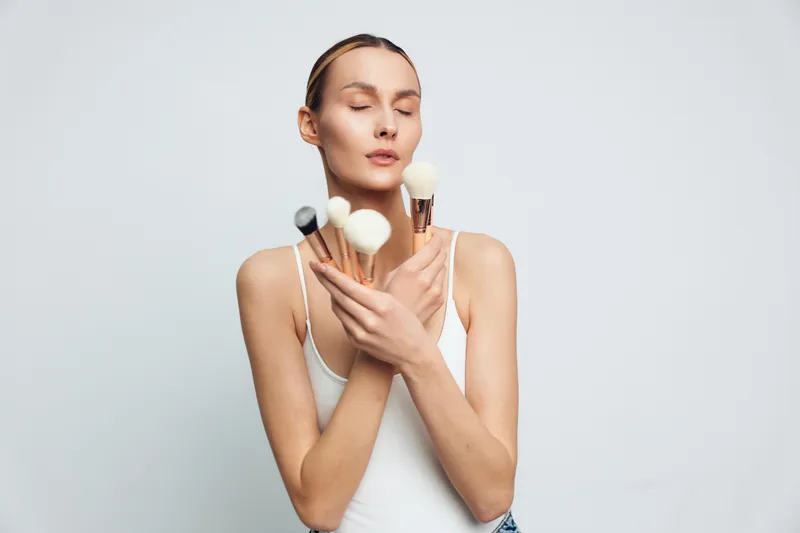 Young woman with closed eyes holding makeup brushes crossed on chest. White background, natural light, concept of beauty, skincare, and self-care routine, calm expression.