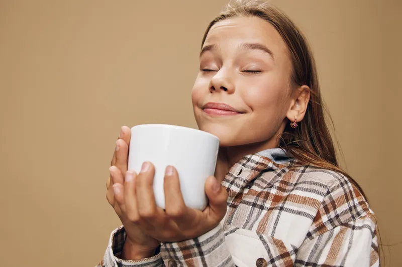 Girl enjoying warm drink with closed eyes and smile wearing casual plaid shirt holding white cup on beige background.