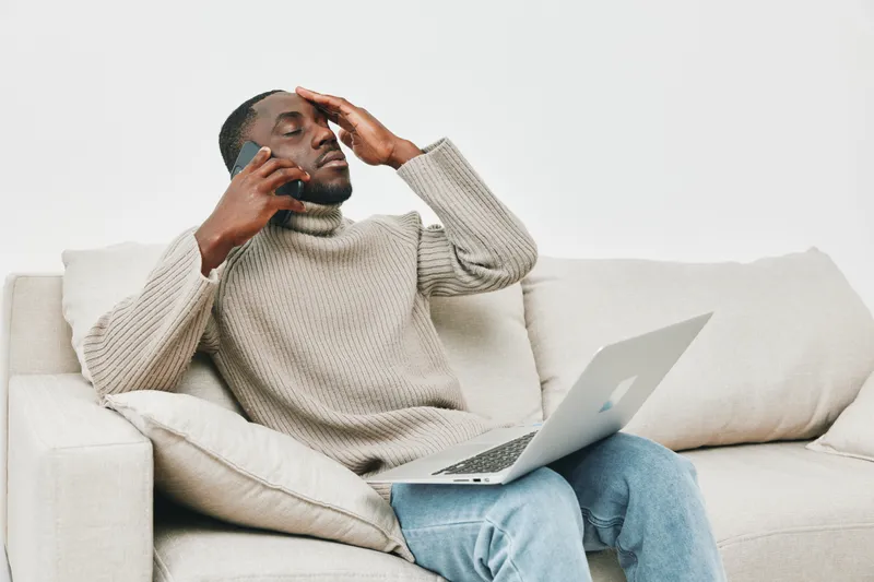 Stressed young African man working remotely on a laptop, feeling overwhelmed while talking on the phone, wearing a cozy sweater in a bright living room