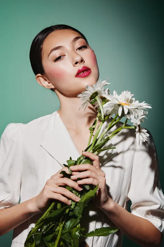 Young woman with radiant skin holds white daisies close wearing fashionable white blazer in studio on green background for beauty creative fashion concept.