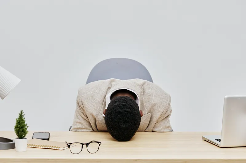 Overworked man with head on desk in office workspace, surrounded by laptop, eyeglasses, smartphone, and plant, demonstrating stress and fatigue. Modern workplace stress concept.