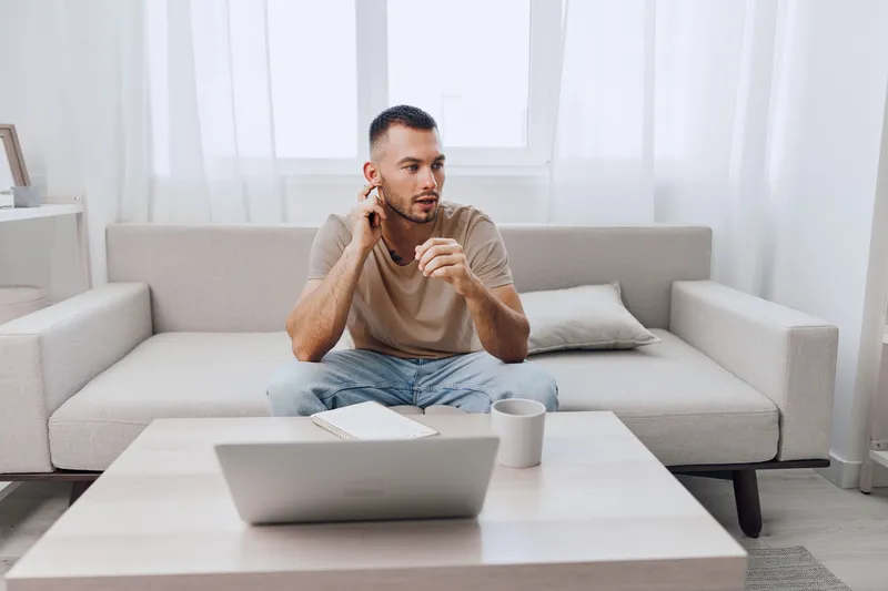 Thoughtful man sitting on a couch, engaged in a conversation while working from home. Comfortably dressed, he expresses concentration and creativity in a modern setting.