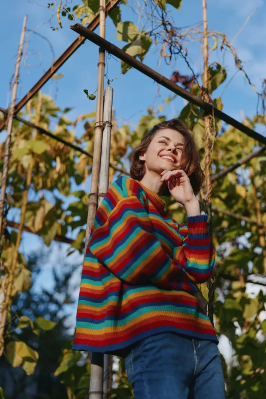 Woman in a colorful striped sweater leaning on a bamboo trellis outdoors, smiling and thoughtful in a casual outfit with jeans in an autumn garden lifestyle and fashion portrait.