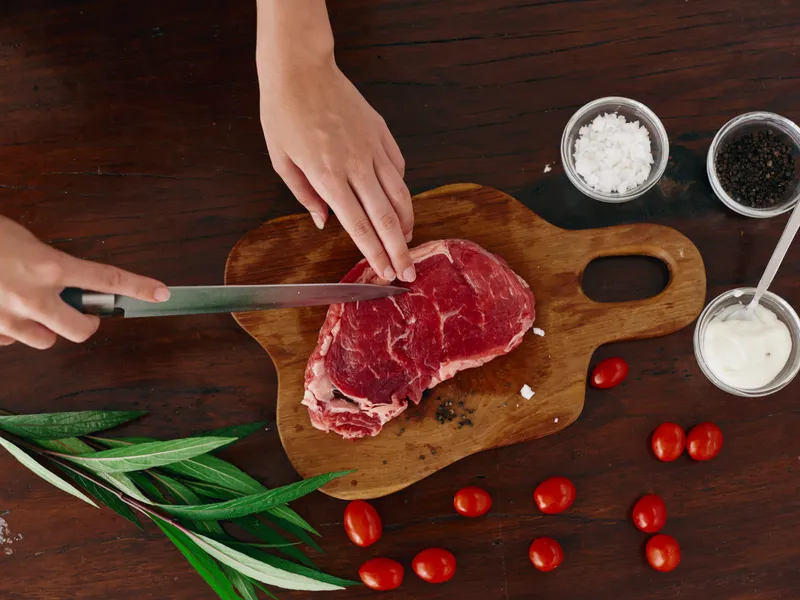 Женщина с ножом в руке нарезает свежее стейковое мясо для жарки на кухне