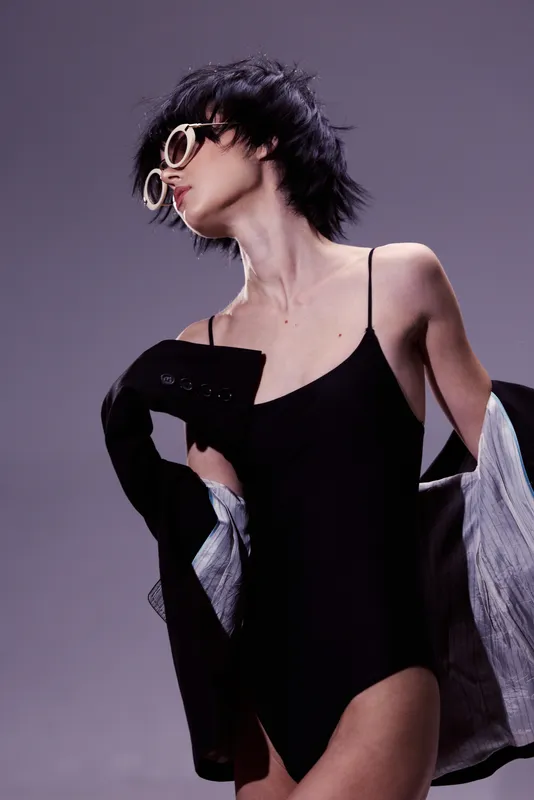 Stylish woman posing in black bodysuit with sunglasses and jacket on gray background, fashion, portrait, trendy, confident