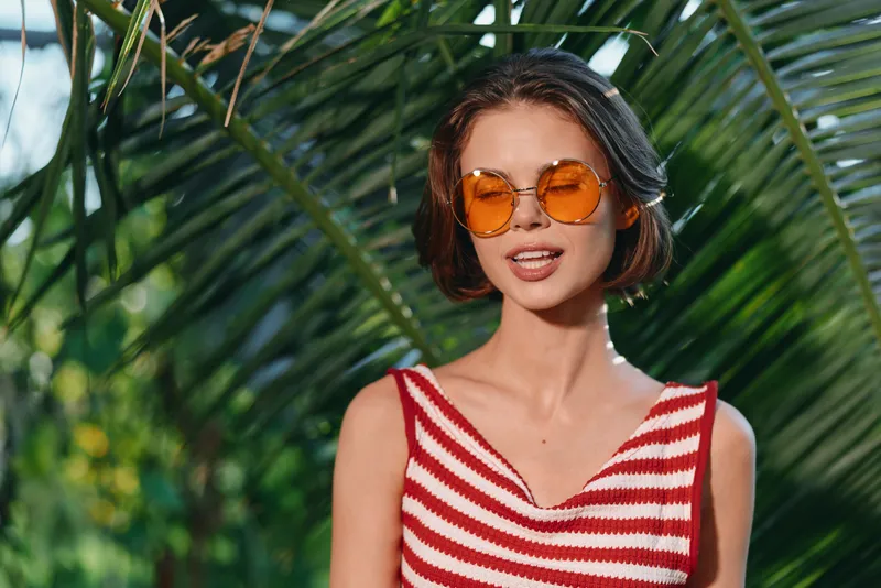 A cheerful woman enjoys a sunny day outdoors in a vibrant striped top and tinted orange sunglasses. She smiles softly, radiating relaxed confidence and casual style.