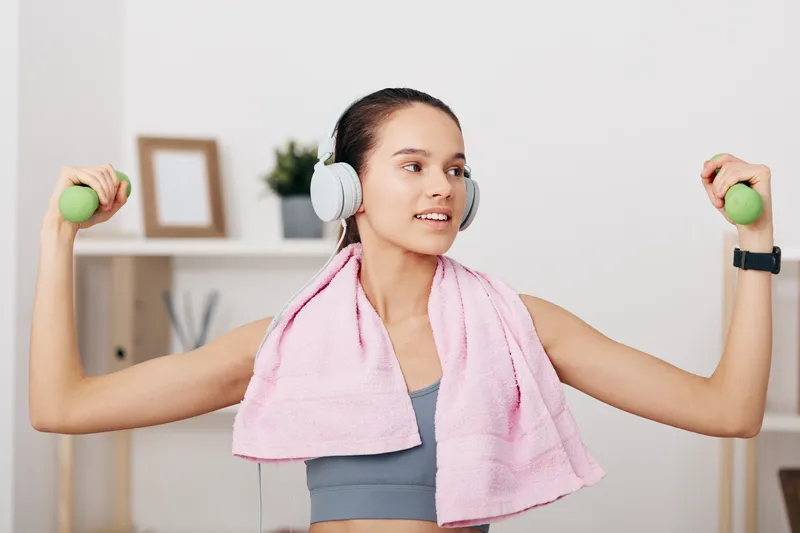 Fitness woman in a home gym, lifting light green dumbbells with a pink towel over shoulders and headphones, ready for workout and focus on strength and cardio.