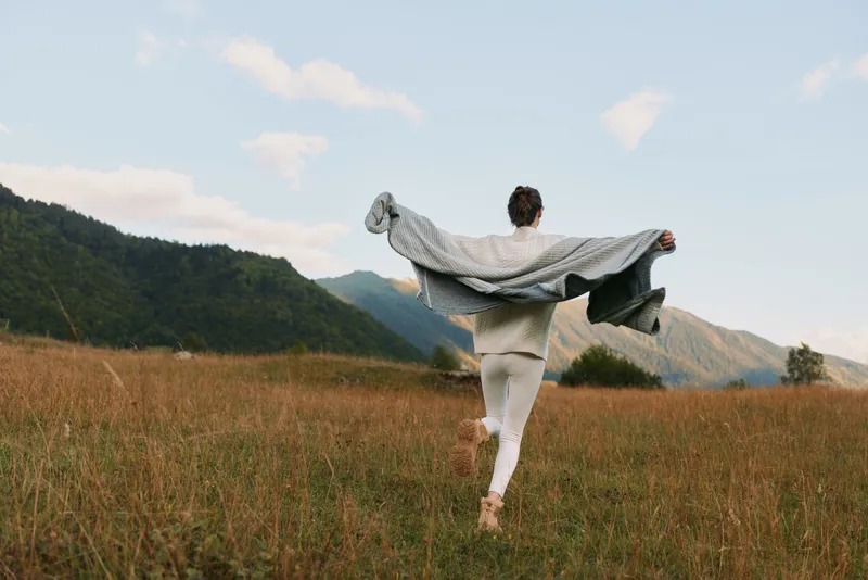 A woman in casual clothing runs through a grassy field with mountains in the background, holding a blanket open behind her. Concept of freedom, nature, and lifestyle.
