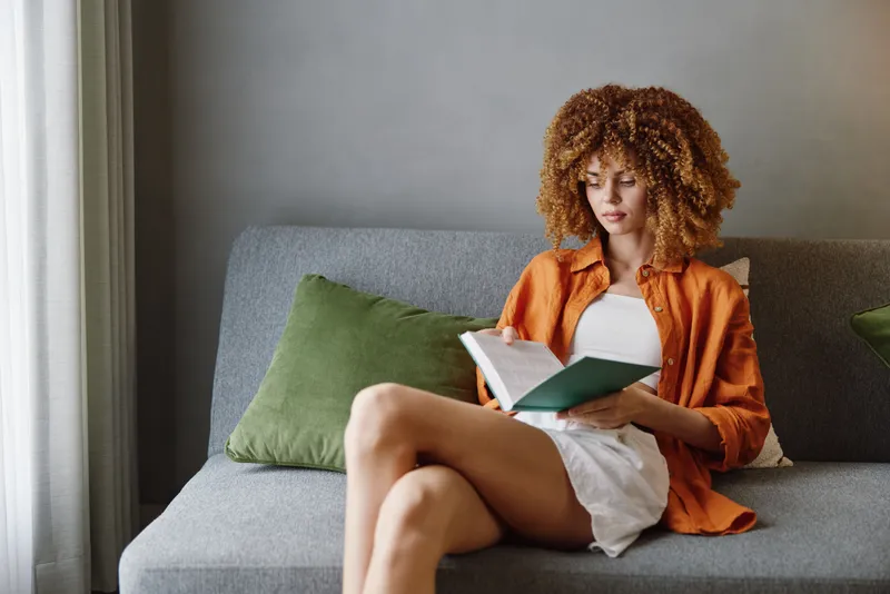 Молодая женщина с вьющимися волосами наслаждается чтением книги на диване в уютной современной обстановке, одетая в оранжевую рубашку и белые шорты, что олицетворяет спокойствие и досуг