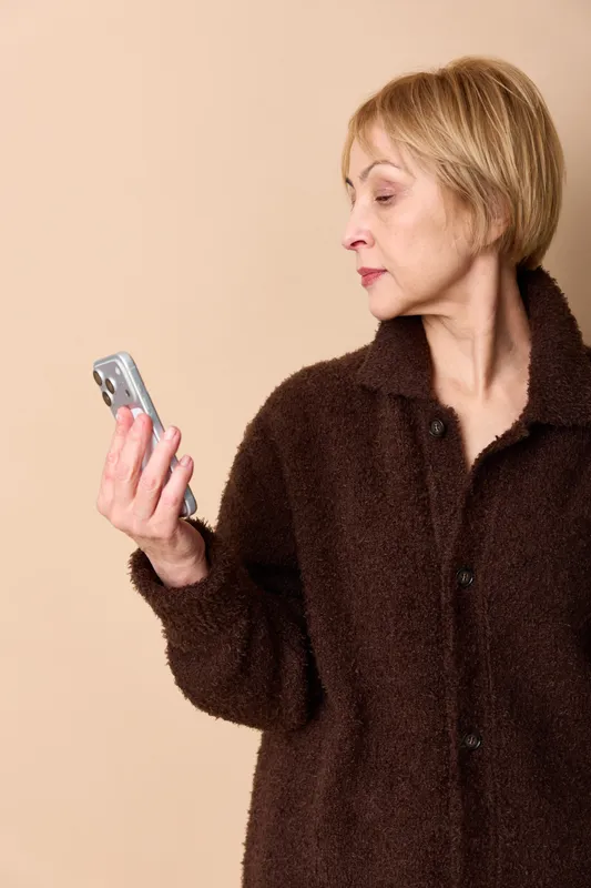 Woman holding smartphone, texting on screen, portrait on beige background, indoors, wearing a robe, focused expression, warm studio light, communication technology concept