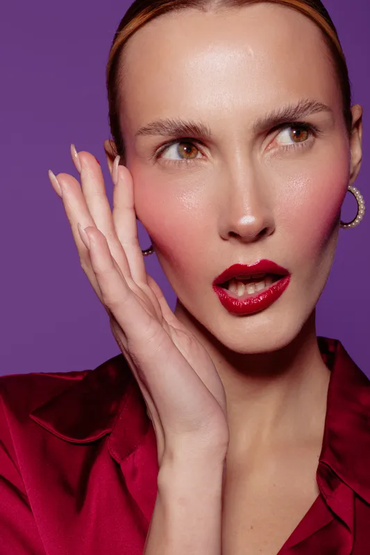 Close-up of a surprised woman with red lipstick and hoop earrings wearing a red satin blouse on purple background. Concept of beauty, fashion, and expressive emotions.