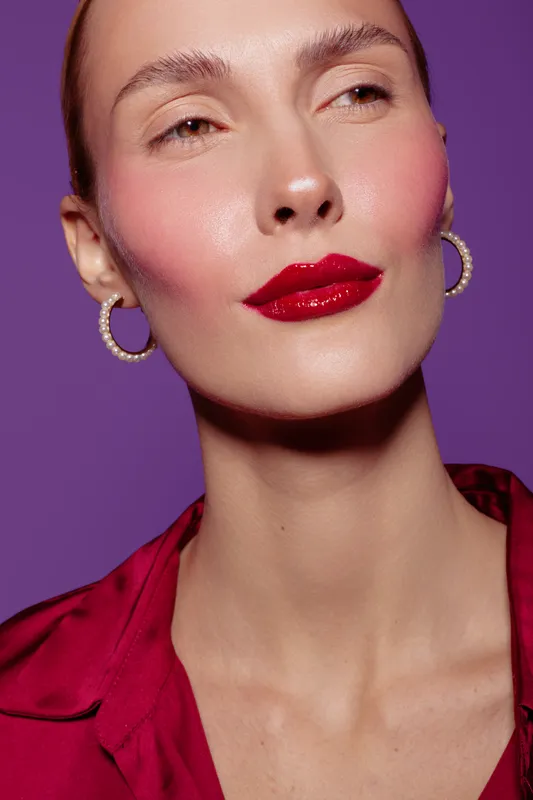 Close-up of a confident woman with glossy red lips and pink blush wearing hoop earrings and a red blouse. Purple background highlights makeup and fashion beauty concept.