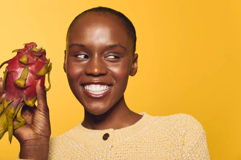 Smiling African woman with dark skin holds dragon fruit near face on yellow background. Concept of healthy lifestyle, exotic fruit, and natural beauty with vibrant studio lighting.