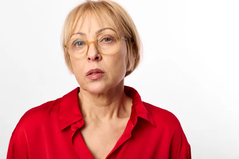 Woman, glasses, red shirt, portrait, short blonde hair, mature calm expression, studio background, direct gaze, soft even light, minimal composition