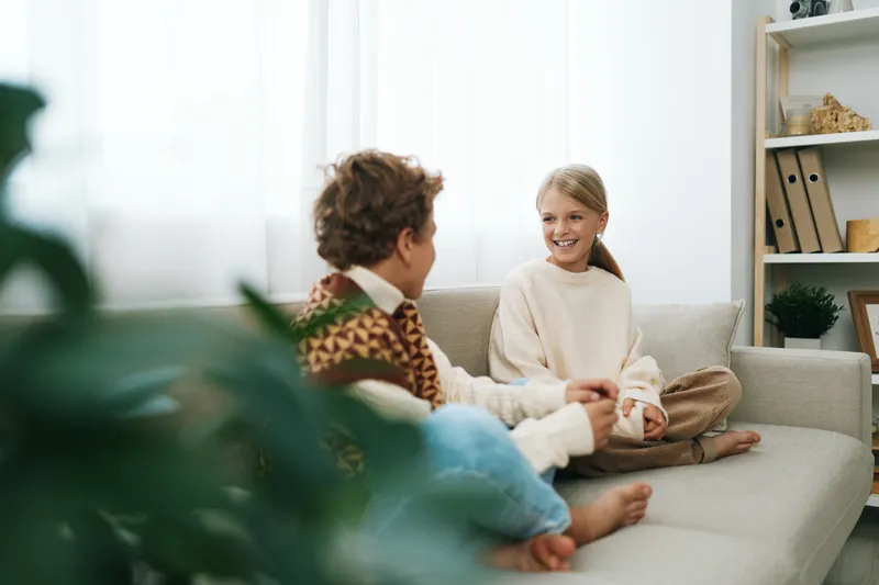 Two children sit on a cozy sofa in a bright living room, sharing a warm conversation with relaxed posture and soft light, creating a candid, inviting moment.