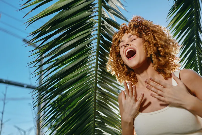 joyful woman with curly hair singing outdoors near tropical palm leaves under clear blue sky, wearing white tank top and expressing happiness.