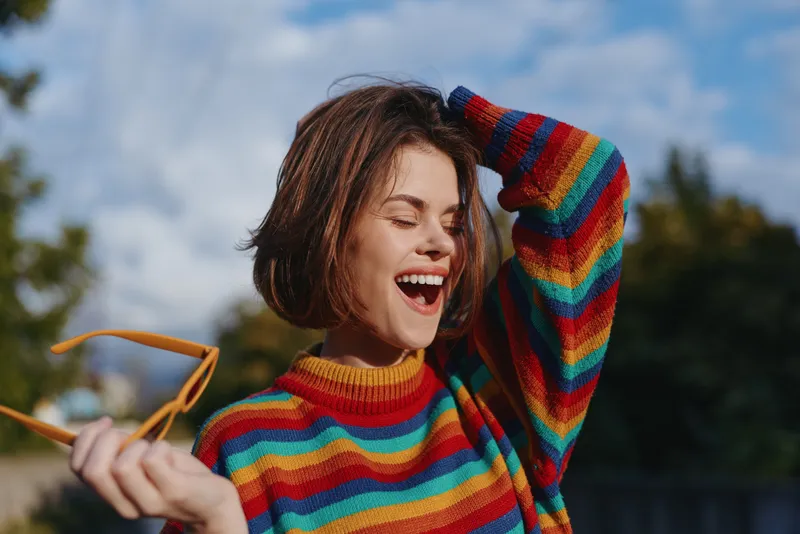 Woman smiling in rainbow sweater holding sunglasses outdoors, joyful portrait of happy young adult with short hair, colorful knit and candid laughter in natural daylight.