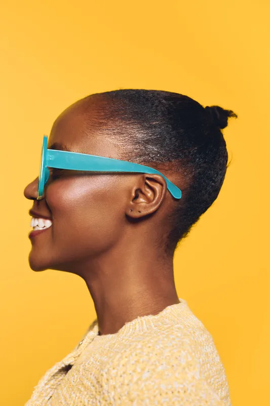 Profile of a smiling dark skinned African woman wearing blue sunglasses and a beige sweater. She stands against a bright yellow background with a cheerful and confident expression.