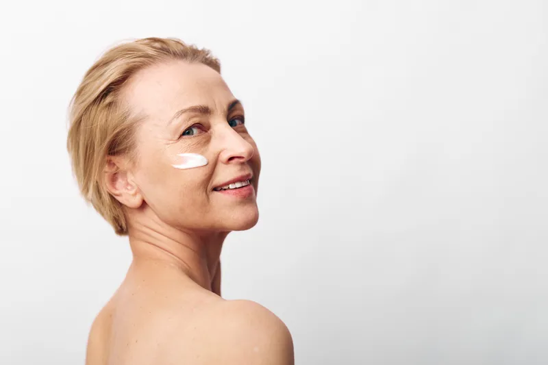 Mature woman with short blonde hair applies white face cream on cheek, smiling gently. Natural skincare concept focused on aging skin care and healthy beauty on plain white background.