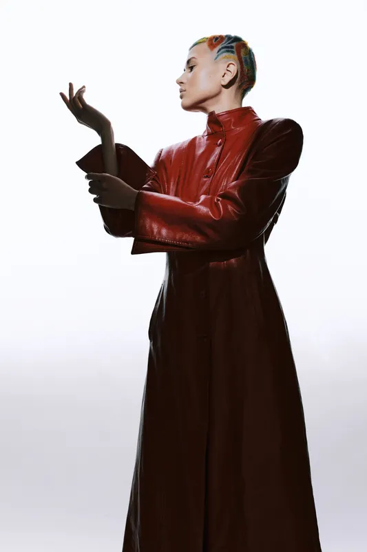 Woman wearing a stylish long red leather coat with colorful braided hair posing gracefully against a clean white background, showcasing fashion and elegance.