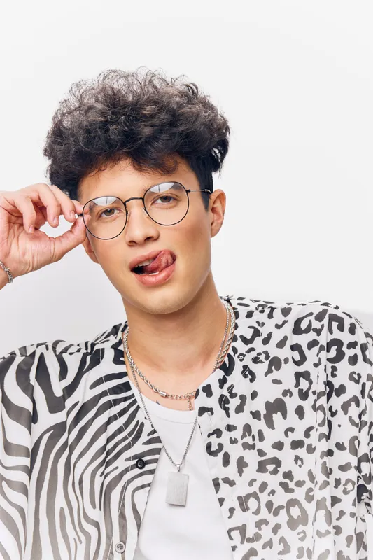 young man with curly hair wearing glasses and animal print shirt making funny face with tongue out against white background