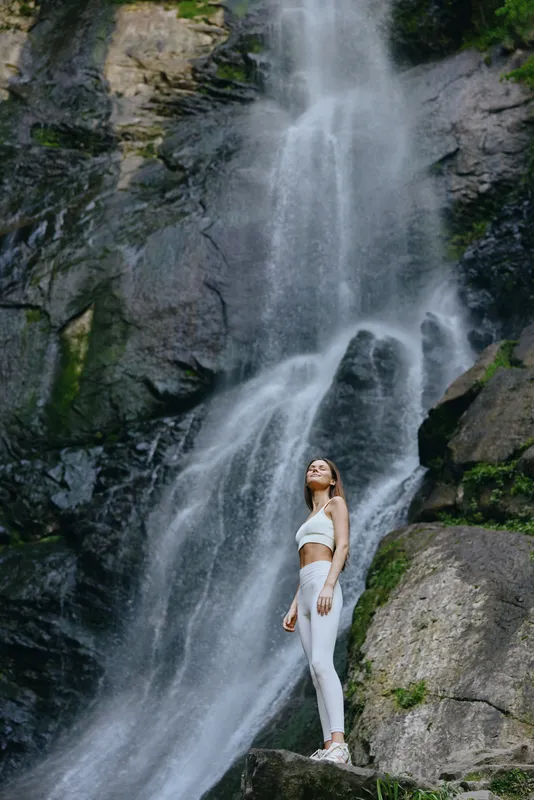 woman standing near waterfall in white activewear, surrounded by rocks and lush greenery, serene and natural outdoor setting with flowing water and mist.