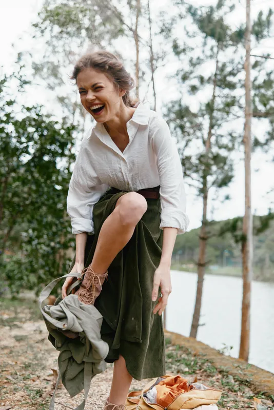 Joyful young woman in a green skirt and white shirt, laughing outdoors by a serene lake, embodying happiness and freedom in nature