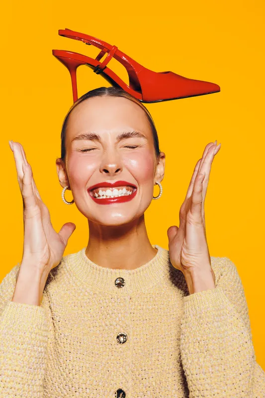 Happy woman with closed eyes and red lipstick smiles widely with red high heel shoe balanced on her head against bright yellow background. Concept of fun, fashion, and joy.