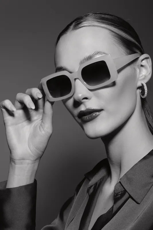 Stylish white woman with fair skin and slicked hair wearing large square sunglasses and hoop earrings in studio. Concept of fashion, elegance, and confidence.