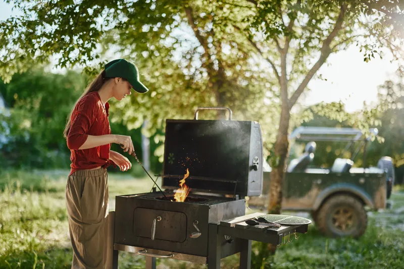 Young woman grilling outdoors in a natural setting during summer, showcasing culinary skills and joy in cooking over a barbecue